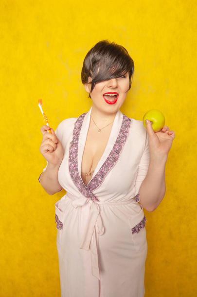 Plump woman with short black hair stands in a pink Bathrobe with teeth brush and green apple on a yellow Studio background alone - Photo, Image