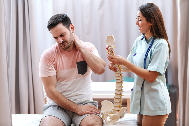 Man in pain sitting on hospital bed and holding neck. Next to him standing doctor and holding model of spine and talking to him. - Photo, Image