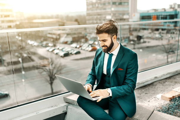 Успешный улыбающийся кавказский бизнесмен в формальной одежде сидит на крыше и пользуется ноутбуком
. - Фото, изображение