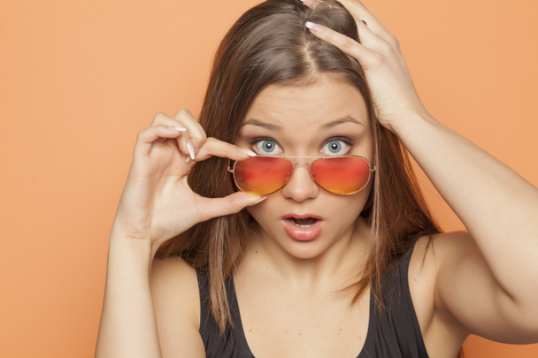 Young startled girl with orange sunglasses on an orange background - Photo, Image