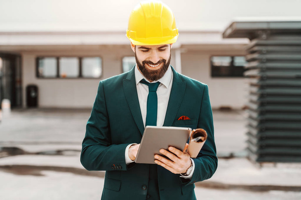 Close up of successful architect in formal wear and yellow helmet using tablet. Under armpit plans. - Foto, Imagen