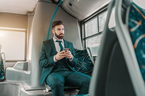 Caucasian businessman in formal wear and serious facial expression sitting in public transportation and using smart phone. - Φωτογραφία, εικόνα