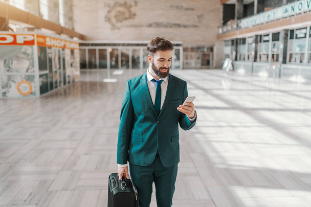 Dreiviertellänge eines lächelnden bärtigen Geschäftsmannes in formeller Kleidung, der sein Gepäck in der Hand hält und während er am Flughafen steht, sein Smartphone benutzt. Geschäftsreisekonzept. - Foto, Bild