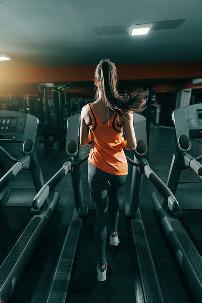 Slim brunette in orange sportswear and ponytail running on treadmill in gym at night. Backs turned. Do it now, sometimes later becomes never. - Photo, Image