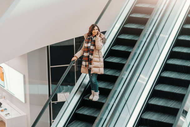 Top view of smiling brunette in jacket and with scarf talking on the phone while riding escalator. Shopping mall interior. - Photo, Image