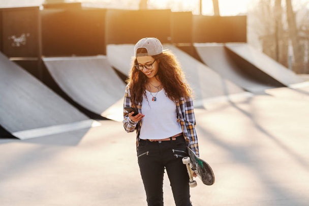 Flaco mixta raza adolescente urbana chica usando el teléfono inteligente y la celebración de monopatín, mientras que de pie en el parque de skate
. - Foto, Imagen