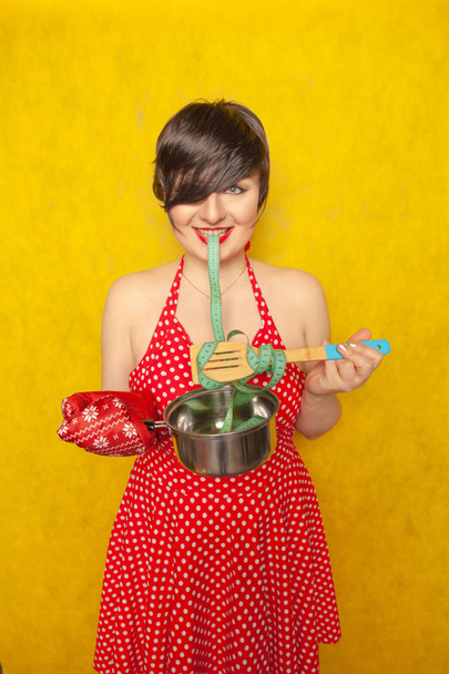 mollig meisje in een rode polka-dot jurk staat met een lege pan en een centimeter tape op een lepel, wil eten, maar de diëten voor gezondheid en figuur - Foto, afbeelding