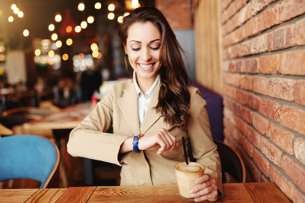 Приваблива брюнетка з зубною посмішкою одягнена в розумний випадковий погляд на наручний годинник і пити каву в кафетерії
. - Фото, зображення