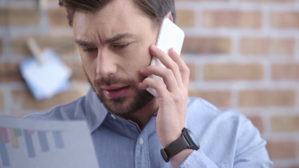 Бизнесмен в синей рубашке с наручными часами разговаривает на смартфоне и смотрит на документ на рабочем месте
 - Кадры, видео