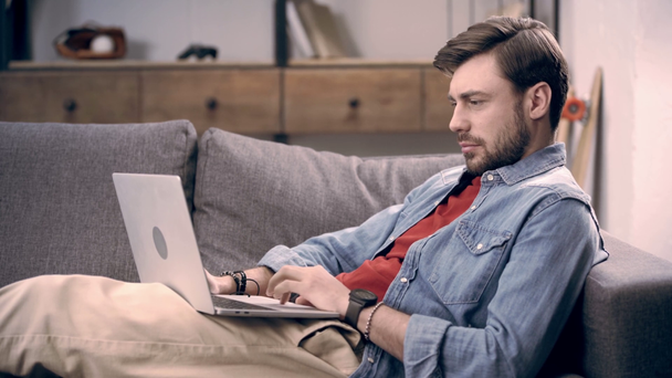Freelancer in denim shirt lying on couch with laptop, typing on keyboard and blinking while looking at screen - Footage, Video