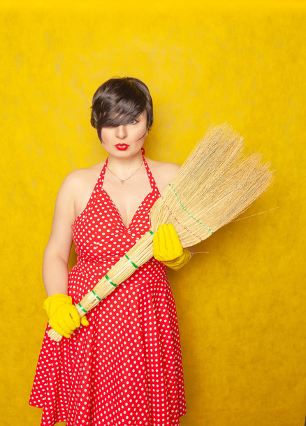 スタジオで黄色の固体背景に宿題手にほうきで掃除用のゴム手袋で赤いレトロ水玉ドレスでかわいい魅力的な感情的な若い女性が立っています。 - 写真・画像