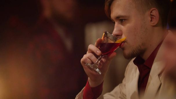 Привлекательный мужчина нюхает и пьет красный ликер в баре с мягким внутренним освещением
. - Фото, изображение