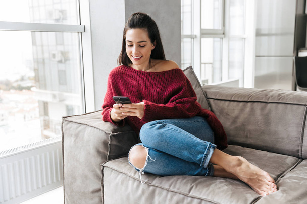 Beautiful woman indoors in home on sofa using mobile phone. - Foto, Imagen