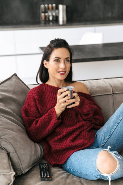 Woman indoors in home on sofa watch TV drinking coffee. - Fotoğraf, Görsel