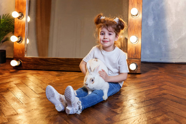 Little girl sitting on floor and playing with white rabbit bunny in modern room over mirror - Fotografie, Obrázek
