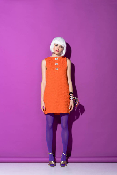 Gorgeous girl in white wig and orange dress standing on purple background - Foto, imagen