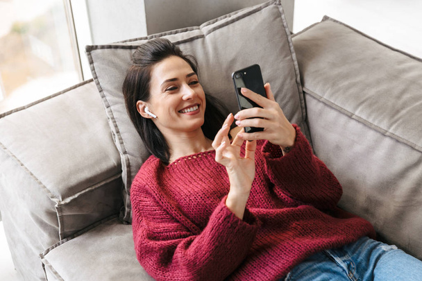 Beautiful woman indoors in home on sofa using mobile phone. - Foto, imagen