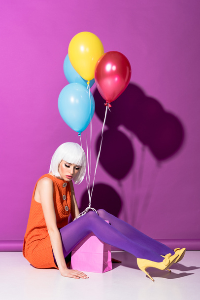 Sad young woman in white wig with air balloons sitting on purple background - Photo, Image
