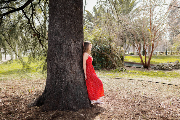 Jeune femme blonde souriante avec robe rouge appuyée contre un arbre
 - Photo, image