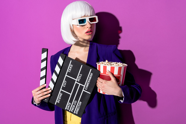 Worried girl in 3d glasses holding clapperboard and popcorn on purple background - Φωτογραφία, εικόνα