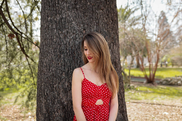 Jovem mulher loira sorridente com vestido vermelho encostado a uma árvore
 - Foto, Imagem