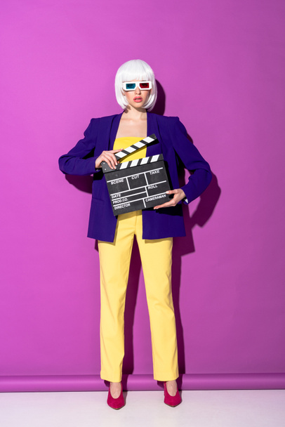Stylish young woman in 3d glasses holding clapperboard on purple background - Фото, изображение