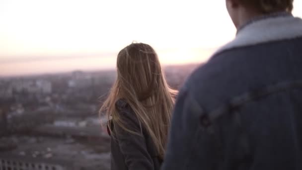 Millenial stylish couple - long haired girl and blondy man standing on the windy roof embracing with a beautiful scene of sunrising on the background. Blurred cityscape around - Πλάνα, βίντεο