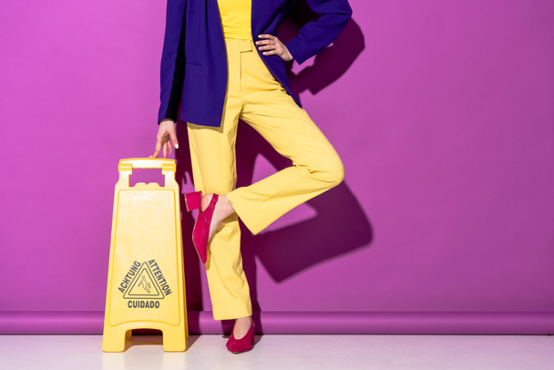 Cropped view of stylish woman standing on one leg near wet floor sign on purple background - Photo, Image