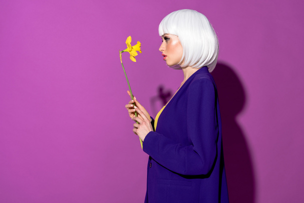 Side view of elegant girl in wig holding flower on purple background - Zdjęcie, obraz