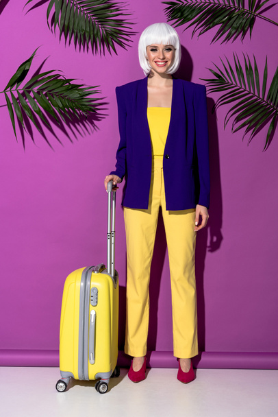 Full length view of smiling girl in wig with yellow suitcase standing on purple background - Fotoğraf, Görsel