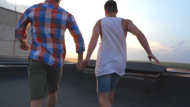 Friends rejoices life during standing on roof at sunset time. Gay couple raising hands showing joyful emotions. Men standing rooftop and victoriously outstretching arms up. Beautiful view. Slow motion - Footage, Video