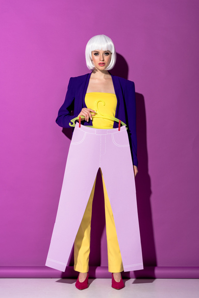 Full length view of woman in white wig holding hanger with paper pants on purple background - Photo, Image