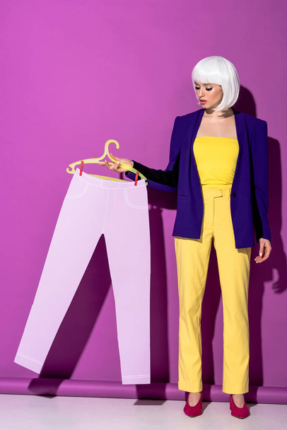 Full length view of elegant girl in white wig holding hanger with paper pants on purple background - Фото, изображение