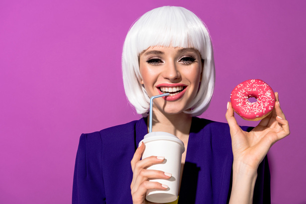 Smiling woman in wig drinking beverage and holding doughnut on purple background - Φωτογραφία, εικόνα