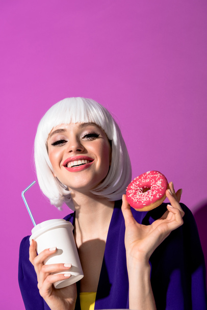 Cheerful girl in wig holding beverage and doughnut on purple background - Foto, afbeelding