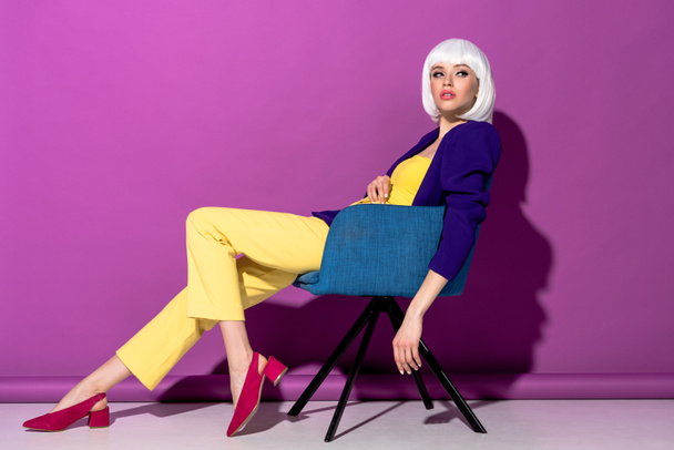 Charming girl in white wig sitting in armchair and looking away on purple background - Foto, Bild