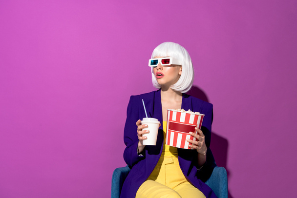 Mujer asombrada en gafas 3d sentada en sillón con soda y palomitas de maíz sobre fondo morado
 - Foto, Imagen