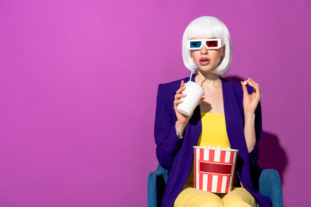 Stylish woman in 3d glasses eating popcorn and drinking soda on purple background - Photo, Image
