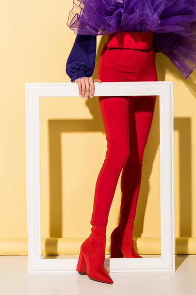 Cropped view of woman in red pants holding white frame on yellow background - Foto, Bild