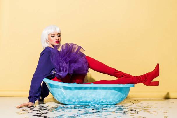 Stylish young woman in wig lying in blue tub on yellow background - Photo, Image