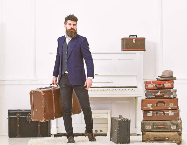 Luggage and vacation concept. Man, traveller with beard and mustache with luggage, luxury white interior background. Macho elegant on strict face stands near pile of vintage suitcase, holds suitcase - Photo, image