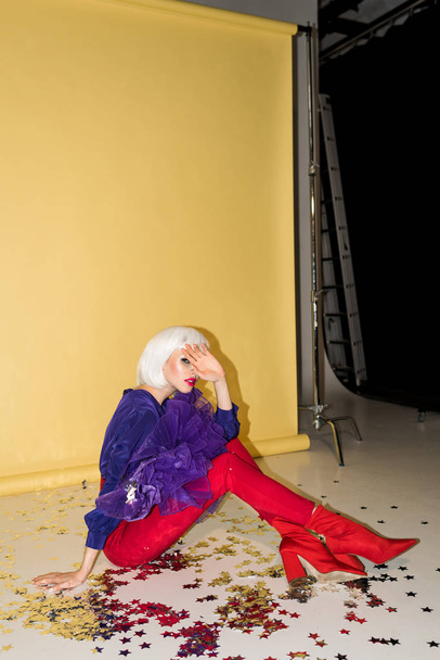 Young woman in wig sitting on confetti and covering face with hand on yellow background - Foto, immagini