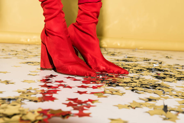 Cropped view of woman in red high-heeled shoes standing on confetti on yellow background - Photo, Image