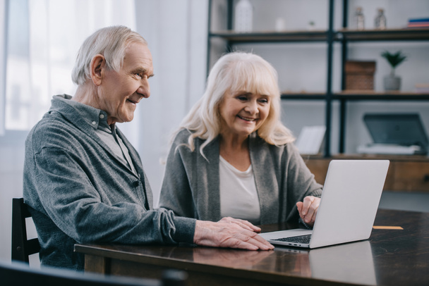 smiling senior couple sitting at table and using laptop at home - Foto, Imagem