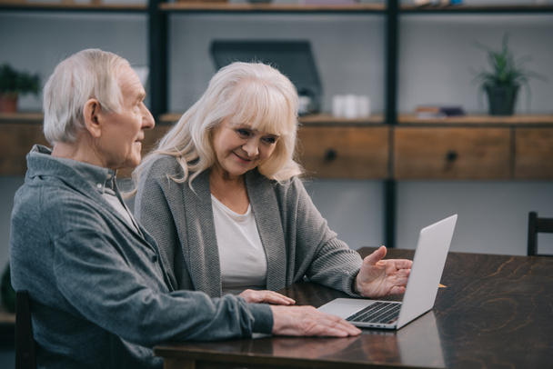 улыбающаяся пожилая пара, сидящая за столом и использующая ноутбук дома
 - Фото, изображение