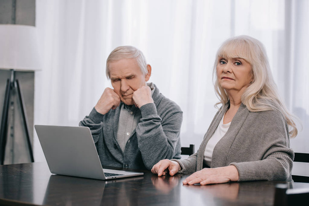 upset senior couple with grey hair sitting at table and using laptop at home - Photo, image