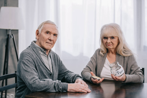 triste pareja de ancianos sentados en la mesa, mirando a la cámara y sosteniendo frasco de vidrio vacío con letras 'pensión'
 - Foto, Imagen