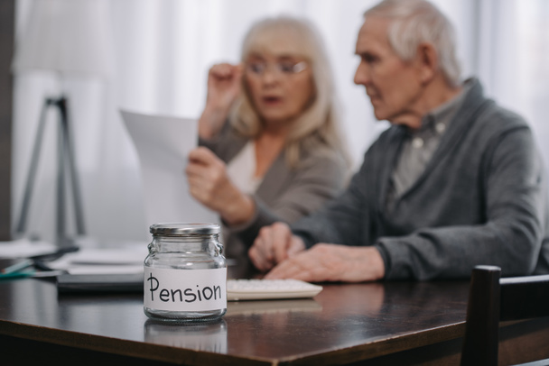 селективный фокус стеклянной банки со словом "пенсия" на столе и пожилой пары на заднем плане
 - Фото, изображение