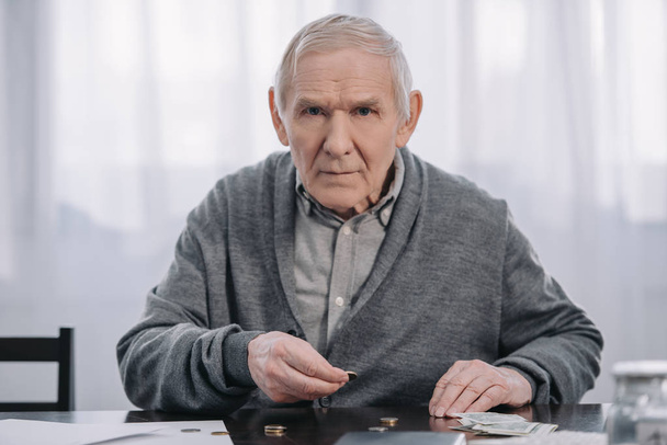 senior man sitting at table, looking at camera and counting money at home - Foto, afbeelding