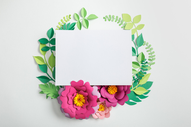 top view of white blank card near pink paper flowers with green leaves on grey background - Photo, Image